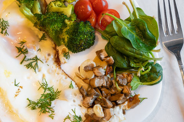 Proper nutrition, healthy eating breakfast of eggs, broccoli, mushrooms, garlic, spinach, dill and small tomatoes close up on white plate with fork lying near. Diet balanced organic food breakfast