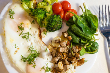 Proper nutrition, healthy eating breakfast of eggs, broccoli, mushrooms, garlic, spinach, dill and small tomatoes close up on white plate with fork lying near. Diet balanced organic food breakfast