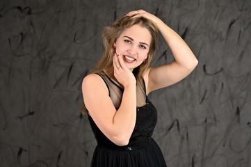 Naklejka premium A photo of a pretty smiling girl with long hair and excellent make-up in a black dress stands in different poses. Universal concept of horizontal female portrait on gray alternative background.