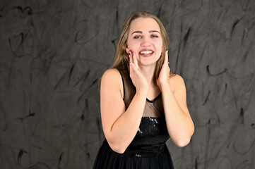 A photo of a pretty smiling girl with long hair and excellent make-up in a black dress stands in different poses. Universal concept of horizontal female portrait on gray alternative background.