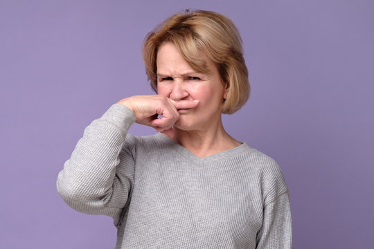 Handsome expressive grandmother coering nose from hodor and bad smell from kitchen. Studio shot. Negative facial human emotion.