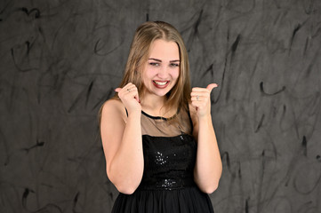 Fototapeta premium A photo of a pretty smiling girl with long hair and excellent make-up in a black dress stands in different poses. Universal concept of horizontal female portrait on gray alternative background.