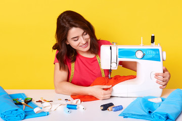 Horizontal picture of hard working professional seamstress sewing item of clothes with help of sewing machine, getting pleasure, smiling sincerely, sitting at table, isolated over yellow background.