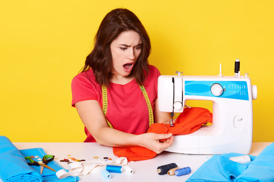 Picture of shocked desperate young inexperienced seamstress working with red fabric, having troubles with sewing, having unpleasant facial expression, opening her mouth widely. Work concept.