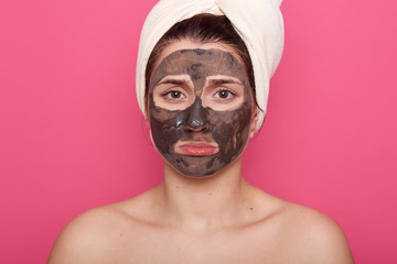 Studio shopt of unhappy beautiful young woman with black clay face mask makes acne therapy, wearing white towel on head, posing against pink studio wall. Beauty, cosmetics and skin care concept.
