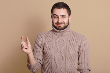 Close up portrait of smiling Caucasian man wearing warm knitted sweater showing small size with fingers, happy bearded European male isolated over beige studio background, demonstrating tiny measure.