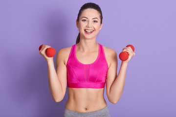 Indoor shot of adorable woman working out with dumbles, looks happy, looking directly at camera...