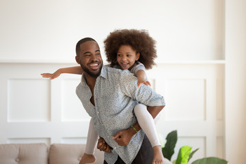 Cheerful african father piggybacking little daughter playing together indoors