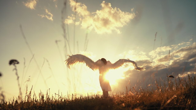 White angel and sun. Brunette girl in a white dress raises angel wings up, looks at the sun