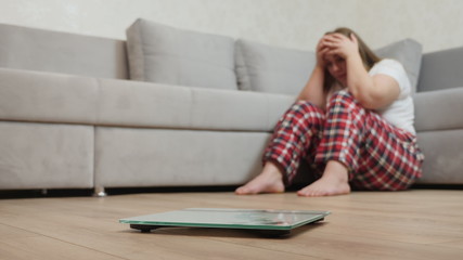 Sad overweight girl with scales on floor. Scale and depressed, frustrated and sad woman sitting on floor holding head and arms on knees