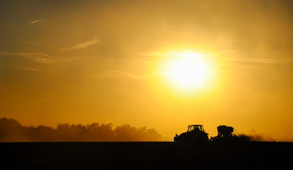 Fototapeta premium Silhouette of a tractor sowing seeds in a field in a cloud of dust against the background of the setting sun.