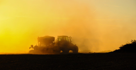 Fototapeta premium Silhouette of a tractor sowing seeds in a field in a cloud of dust against the background of the setting sun.