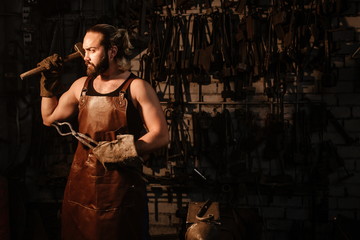 Fototapeta na wymiar Portrait of a brutal muscular blacksmith standing in a dark workshop holding a vise and a hammer