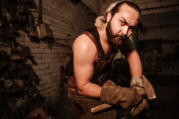 Fototapeta na wymiar Sweaty brutal muscular artisan blacksmith standing in the workshop with a hammer leaning on an anvil