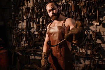 Fototapeta na wymiar Brutal muscular artisan blacksmith standing in the workshop forges iron with a hammer