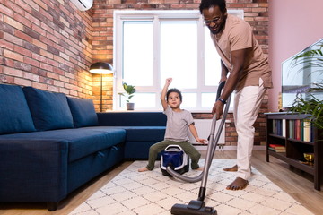 African dad vacuums in the apartment while there is no mom. The child sitting on the vacuum cleaner...