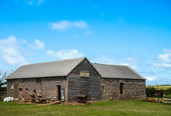 Fototapeta na wymiar Stanley, Tasmania, Australia - December 15, 2009: Hightfield Historic Site. Closeup of gray wooden and stone barn with green lawn under blue sky. Rusty machines in front.