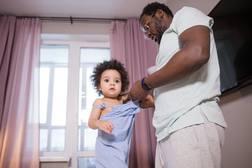 Obraz premium African dad putting clothes on his daughter standing in the bedroom