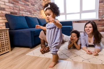 Happy ethnic family lying on the floor watching their daughter learn to walk taking the first steps