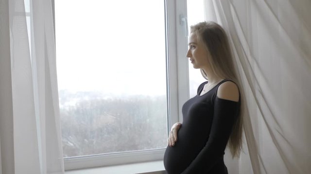 Young pregnant woman standing by the window and holding her belly