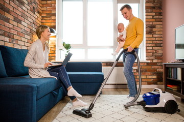 Gender equality. Dad with a baby in his arms, vacuuming garbage with a vacuum cleaner. Mom on maternity leave is sitting on the couch making money online on her laptop