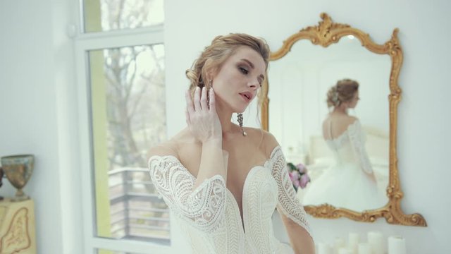 Blonde bride in white wedding dress poses in front of camera, turns seductively looking at camera. Gently touches the face neck hair head with fingers. Behind large stylish mirror with candles flowers