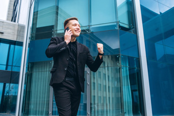The guy in the black suit on the background of the glass modern business center. confident businessman talking on phone walking in front of modern architecture. Portrait of a smiling businessman