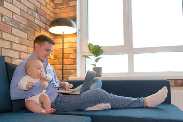 Dad on maternity leave holding a newborn baby sitting on the couch working on a laptop. Gender equality. Dad and child freelancer