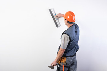 The builder smoothes the wall with a spatula. In work clothes and hard hat. On a light gray background.