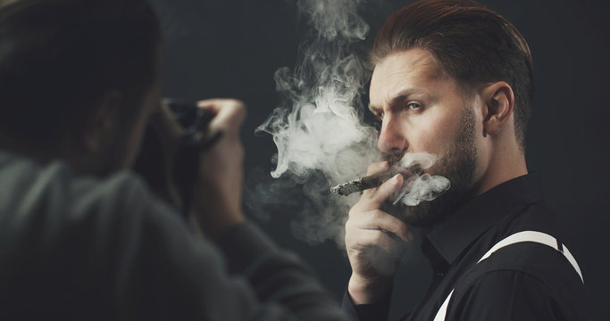 Photographer with camera shooting adult man smoking cigar over dark background, cropped shot