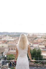 Fototapeta na wymiar Sexy girl in a white dress and blond hair admires Rome in Italy standing on the balcony and looking at historical buildings. Tourist vacation.