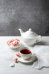 Beautiful romantic breakfast with red tea Hibiscus and meringue cookies in the shape of hearts and candy. White tea-set stand on a wooden table with a concrete background. Vertical with copy space.