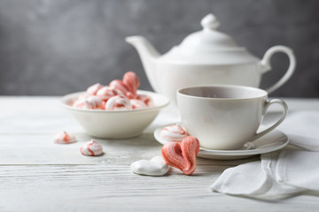 Fototapeta na wymiar Romantic breakfast for a Valentines Day with tea and meringue cookies in the shape of hearts and candys. White tea-set stand on a wooden table with a concrete background. Close up still life.
