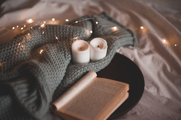Open paper book on wooden tray with knitted sweater with candles over glowing lights closeup in...