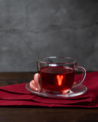 A glass cup of red tea Hibiscus and meringue cookie in the shape of hearts. Stand on textile napkin, wooden table with a concrete background. Still life dark vertical photo minimalism with copy space.