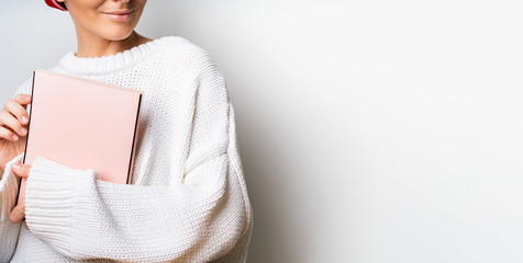 Close view of woman in white woolen sweater holding a book with empty pink cover in hands. Free...