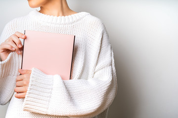 Close view of woman in white woolen sweater holding a book with empty pink cover in hands. Free...