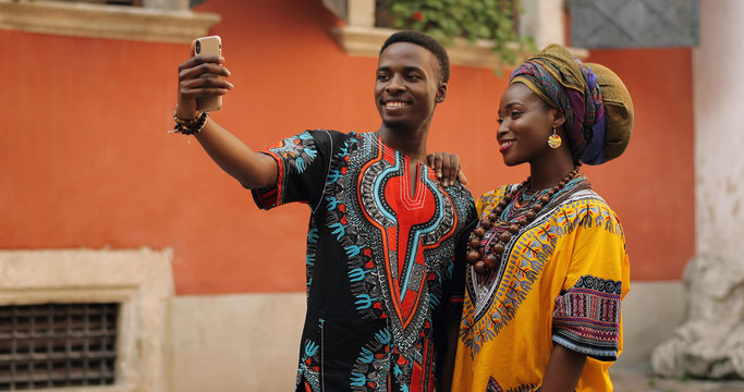 African young stylish and attractive couple in traditional outfits standing together outside and posing to the smartphone camera while taking selfie photo.