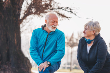 An elderly couple talking while stretching in the park. The woman is wearing blue headphones.