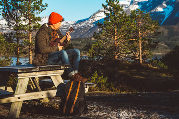 Young man playing guitar sitting on a wooden table against the background of mountains, forests and lakes. Relaxing and enjoying sunny days. Place for text or advertising