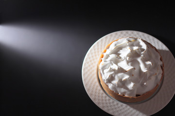 A cake with merengue frosting on a white plate. Black background. Hard light from the left. Space for text