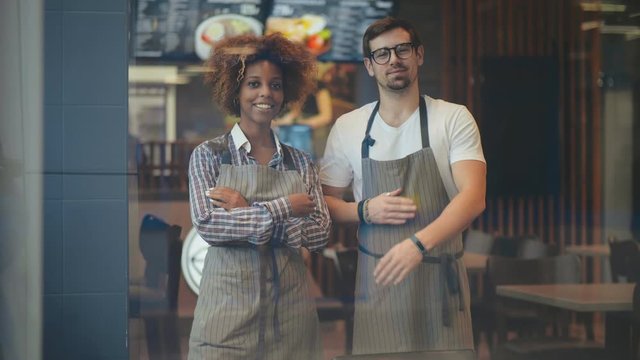 Portrait of multicultural smiling couple of waiters standing in cafe and looking at camera