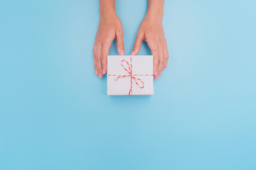 Female hands are holding a white gift box with a thin ribbon as a present for Christmas, New Year, Mother's Day or anniversary on a blue table background, top view. Place for text.