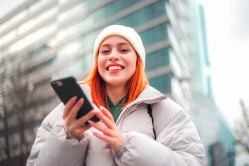 Attractive young redhead woman typing a message via her mobile phone in a city, she is smiling