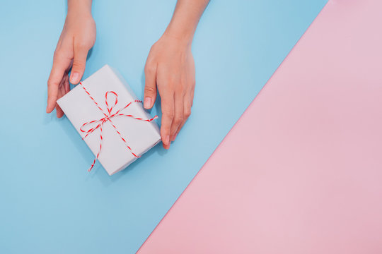 Female hands hold a white gift box with a thin ribbon as a present for Christmas, New Year, Mother's Day or anniversary on a diagonal blue and pink table background, top view. Place for text.