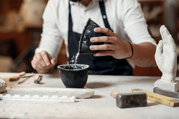 Unrecognizable professional stonemason with well-maintained hands making glue in special vessel for his handmade sculpture - obrazy, fototapety, plakaty