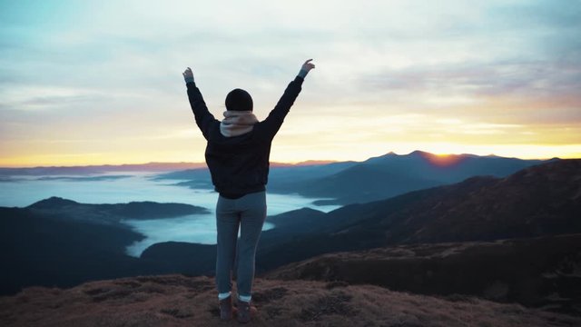 Back view of unrecognizable woman raises his hads up while standing on the mountain top, enjoying the moment and observing the distant hills forest harmony relaxation happiness amaizing view sunrise