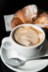 Authentic steaming hot cappuccino in modern white coffee cup next to a fresh croissant in an Italian cafe in Rome, Italy