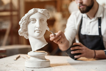Skillful sculptor makes professional restauration of gypsum sculpture of woman's head at the creative workshop. - obrazy, fototapety, plakaty