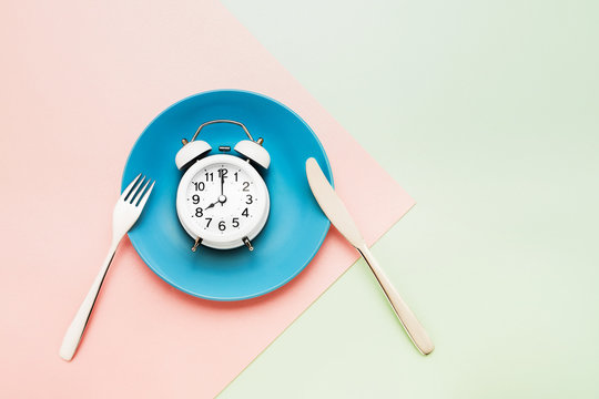 Intermittent fasting concept. White alarm clock on empty blue dish with knife and fork on pink-green background. top view, copy space.
